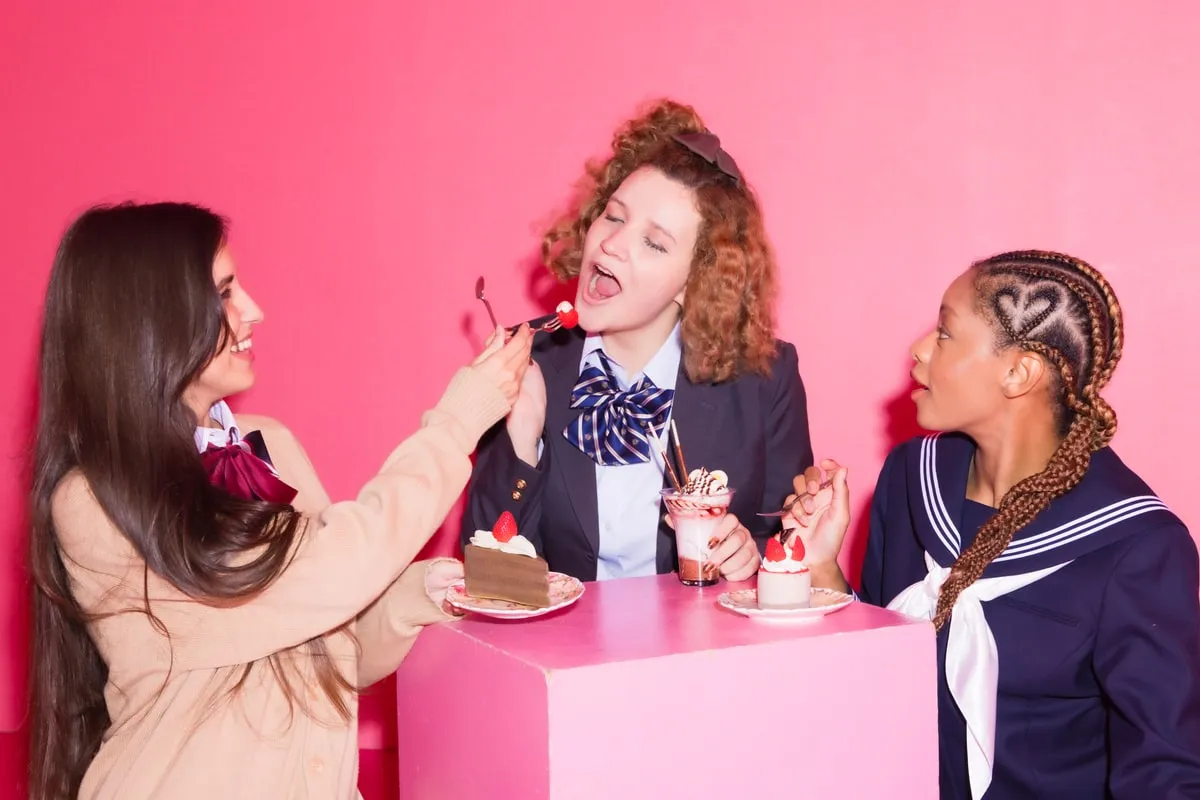ピンクの背景で制服姿の3人がスイーツを食べさせ合う